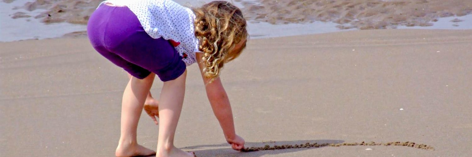 A small girl is bent over on a beach, drawing a line in the sand
