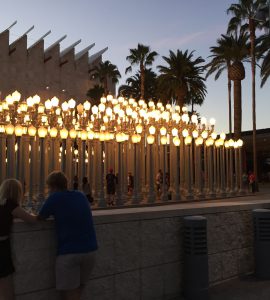 The Stanley Kubrick exhibit at Los Angeles County Museum of Art. It looks like a series of ornate, lit street lamps with people walking among them. There are palm trees, and two people in the foreground talking. It is dusk.
