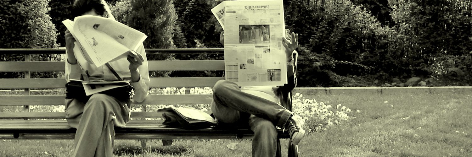 [IMAGE] Two people sat on a part bench reading newspapers. Their faces are obscured.