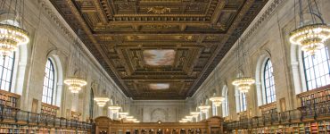 [IMAGE] The New York Public Library main branch reading room. People seated at large desks under a painted ceiling with window lined walls
