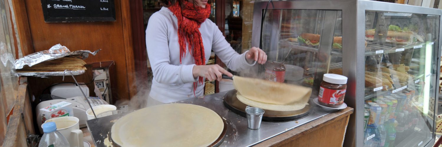 [IMAGE] A light-skinned woman who appears to be in her 20s, wearing a white sweater and bright red scarf, prepares crepes outside of Quasimodo Cafe on the Rue D'arc in Paris