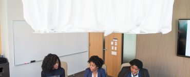 [IMAGE] Three women of color sat next to each other at a table in a meeting room, engaged in conversation. The camera is positioned high above them, looking down