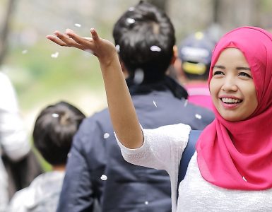 [Image] A woman wearing red hijab smiling and throwing blossom into the air. In the background out of focus are several people not looking at her