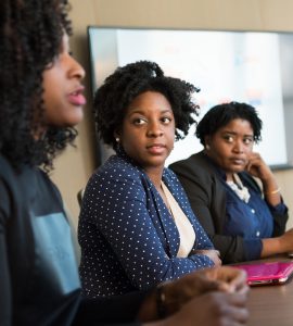 Three black women are seated at a table with computers in front of them. One is speaking, the others are listening.