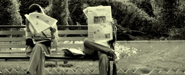 [IMAGE] Two people sat on a part bench reading newspapers. Their faces are obscured.