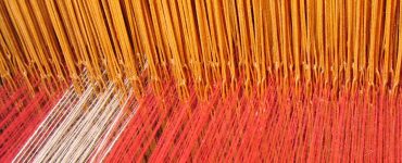 [IMAGE] A close-up of a loom with red, yellow, and white threads combining together.