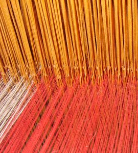 [IMAGE] A close-up of a loom with red, yellow, and white threads combining together.