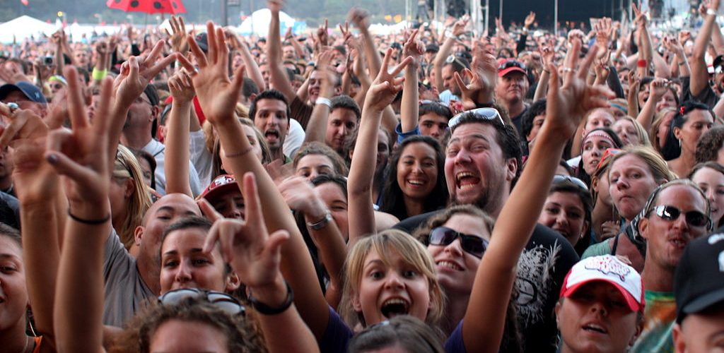 [IMAGE] A large crowd of people with arms raised, smiling at the camera. In the background is an umbrella that looks like a ladybug. The image appears to have been taken of a crowd at a concert during the day time.
