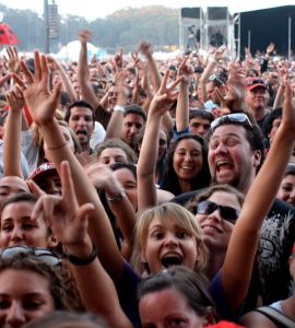 [IMAGE] A large crowd of people with arms raised, smiling at the camera. In the background is an umbrella that looks like a ladybug. The image appears to have been taken of a crowd at a concert during the day time.