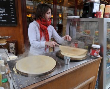 [IMAGE] A light-skinned woman who appears to be in her 20s, wearing a white sweater and bright red scarf, prepares crepes outside of Quasimodo Cafe on the Rue D'arc in Paris