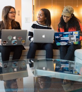 [IMAGE] Three women sat next to each other, talking. All three have laptops on their knees. Some of the laptops have stickers on them.