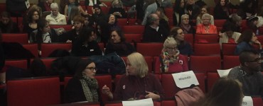 An audience begins to fill in for a panel in an auditorium of red chairs and scattered reserved signs