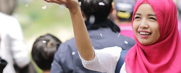 [Image] A woman wearing red hijab smiling and throwing blossom into the air. In the background out of focus are several people not looking at her