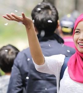 [Image] A woman wearing red hijab smiling and throwing blossom into the air. In the background out of focus are several people not looking at her