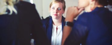 [IMAGE] A blonde women listens to two other women blurred in the foreground with their backs to the camera. They are dressed in professional attire and sat at a table.