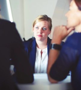 [IMAGE] A blonde women listens to two other women blurred in the foreground with their backs to the camera. They are dressed in professional attire and sat at a table.