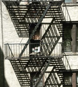 [IMAGE] An outdoor metal fire escape on the side of a building in Manhattan. Shadows from the steps are cast onto the pale brick building behind.