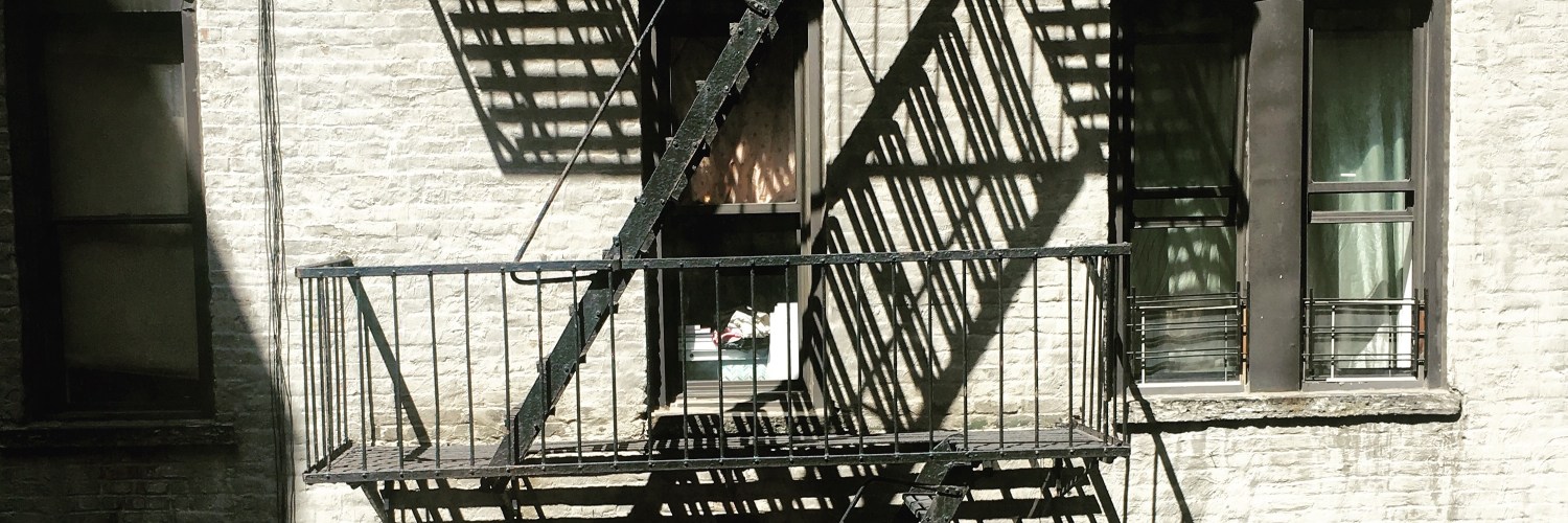 [IMAGE] An outdoor metal fire escape on the side of a building in Manhattan. Shadows from the steps are cast onto the pale brick building behind.