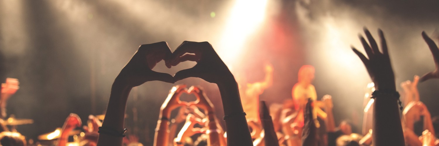 [IMAGE] A crowd at a music concert viewed from behind. A pair of hands make the shape of a heart above the heads of the crowd.