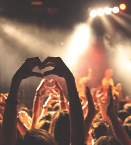 [IMAGE] A crowd at a music concert viewed from behind. A pair of hands make the shape of a heart above the heads of the crowd.